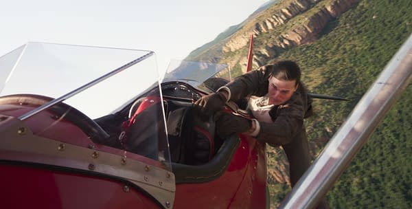 Ethan Hunt, portrayed by Tom Cruise, clings to the side of a red airplane while in flight, showcasing a tense moment from 'Mission: Impossible - The Final Reckoning.' The background features a scenic landscape of green hills and rugged cliffs.