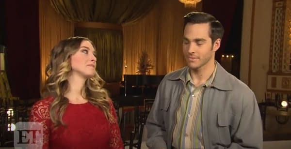 Melissa Benoist, wearing a red dress, and Chris Wood, in a striped shirt, share a moment during an interview. The background features warm lighting and draped curtains, creating a cozy atmosphere.