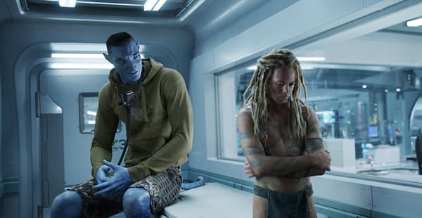 A scene from 'Avatar: Fire and Ash' features Stephen Lang as Quaritch, a blue-skinned Na'vi character dressed in a hoodie, sitting on a bench. Next to him, Jack Champion as Spider, a human character with long, braided hair, sits with his arms crossed, appearing contemplative. The setting is a futuristic environment with screens and technology visible in the background.