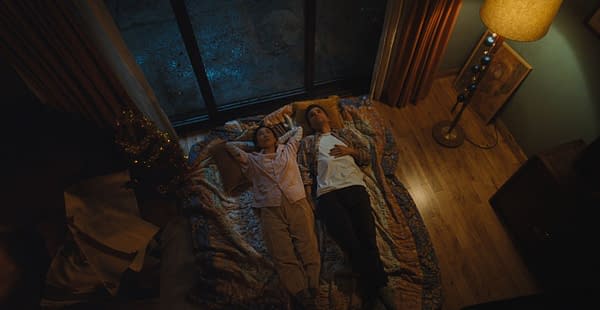 Overhead view of a man and woman lying side by side on a quilt in a dimly lit house, looking up toward the ceiling.