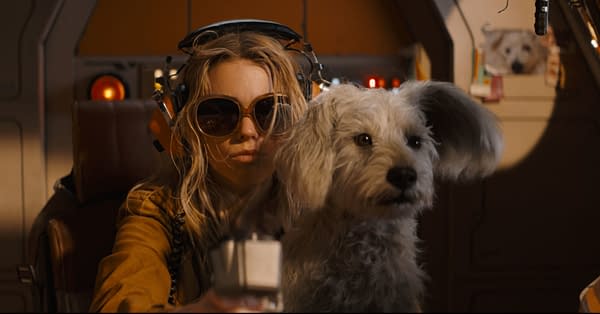 A young woman wearing large sunglasses and headphones sits in a pilot's seat, holding a device and looking serious, accompanied by a fluffy white dog. The interior resembles a futuristic spaceship with warm lighting.