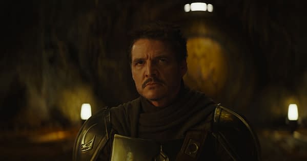 The Mandalorian, a character from Lucasfilm's series, gazes intently in a dimly lit setting, wearing a metallic armor and a dark cloak. The image captures a moment of contemplation in the show's narrative.