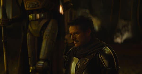 A close-up of the character The Mandalorian, portrayed by Pedro Pascal, wearing a dark helmet and armor, standing near a robot-like figure in a dimly lit environment. This scene is from Lucasfilm's 'The Mandalorian and Grogu.'