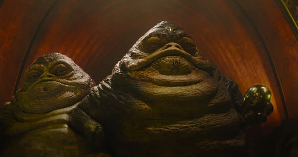 A scene featuring two Hutt characters from Lucasfilm's 'The Mandalorian and Grogu.' The characters have large, rounded bodies and distinctive facial features, set against a warm, wooden background.