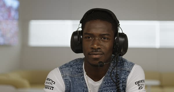 F1: A person with short hair wearing a headset and a denim vest, looking directly at the camera with a calm expression. The background features blurred screens, suggesting a motorsport environment.