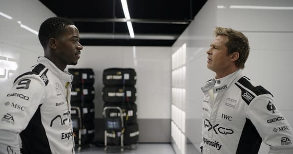 Two race car drivers, one Black and one white, stand facing each other in a well-lit garage, wearing matching racing suits. Behind them are stacked tires and a technical display, setting the scene for a competitive motorsport environment.