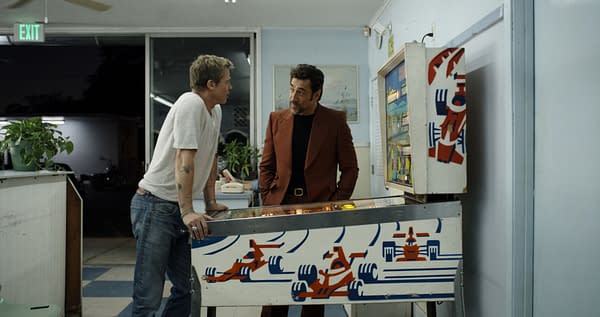 F1: Two men converse in a retro diner setting, leaning over a pinball machine featuring a colorful racing car design. One man is dressed casually in a gray t-shirt and jeans, while the other wears a stylish brown suit.