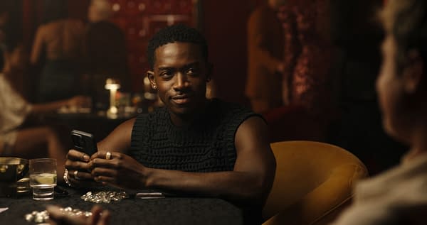 A man with short curly hair and a sleeveless black top sits at a table in a lively setting, holding a smartphone. The background includes blurred figures and illuminated decorations, suggesting a social or nightlife atmosphere.