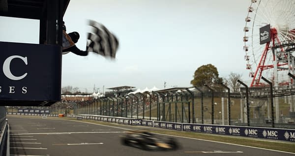 A race official waves a checkered flag from a pit tower at a Formula 1 race, with a blurred car speeding past and a Ferris wheel visible in the background.