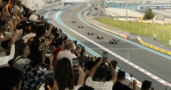 F1: A crowd of enthusiastic Formula 1 fans captures the excitement of a race on their smartphones, as several cars compete on a winding racetrack. The vibrant atmosphere is heightened by the spectators' cheers and the colorful team apparel.