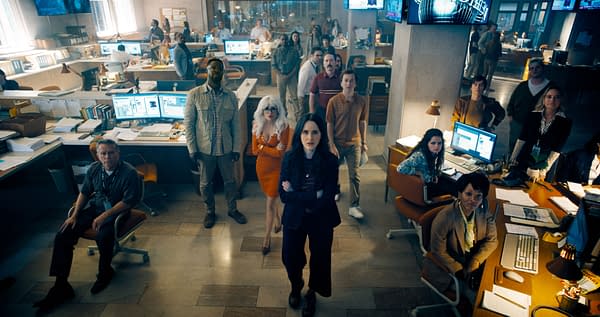 A scene from the film 'Superman' featuring a group of diverse characters in an office setting, with several people gathered around computer desks, showing expressions of concern and focus as they watch something on screens.