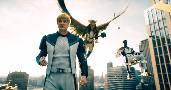 A scene from a superhero film featuring a main character in a futuristic outfit with a glowing green object, flanked by two flying characters, one with bird-like wings and the other seated in a high-tech chair, set against a city skyline.