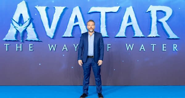 A man in a blue suit stands in front of a large backdrop featuring the title "AVATAR: THE WAY OF WATER" at a premiere event. The background is a gradient of blue and purple colors.