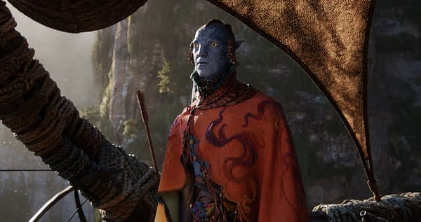 A character named Peylak, portrayed by David Thewlis, is depicted in a vibrant orange cape with intricate designs, set against a coastal backdrop in a scene from 'Avatar: Fire and Ash'. The character has blue skin and striking yellow eyes, embodying a distinct visual style characteristic of the film.