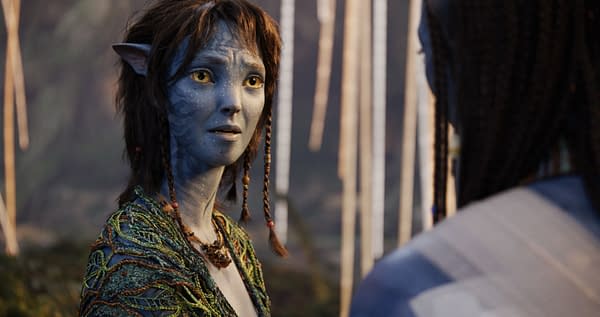 Kiri, a character from 20th Century Studios' "Avatar: Fire and Ash," is depicted with blue skin, expressive eyes, and braided hair, wearing a colorful costume made of intricate patterns. The background features blurred elements that suggest a lush, fantastical setting.