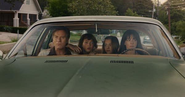 A dramatic scene in a vintage car featuring a man and three children, each displaying varied expressions of concern and intensity while looking out the windshield. The background includes a suburban setting with green trees and houses.