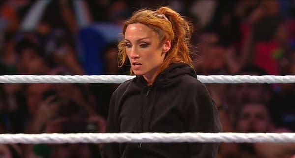 Becky Lynch standing in a wrestling ring at WWE Clash in Paris, visibly focused and surveying the arena, wearing a black hoodie. The backdrop features a cheering crowd.