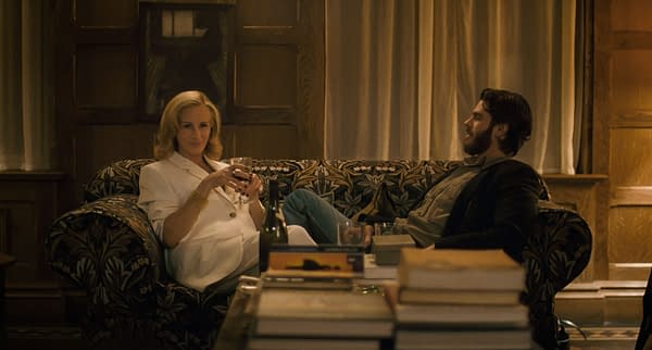 A scene from 'After The Hunt' featuring Julia Roberts as Alma and Andrew Garfield as Hank sitting on a floral couch, engaged in conversation. The setting is cozy, with a coffee table cluttered with books and two wine glasses.