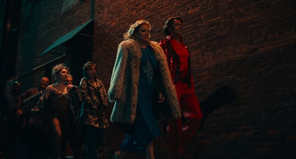 A scene from the film 'Song Sung Blue' shows characters Tom D'Amato, Rachel, Mark Shurilla, Angelina, Claire Stengl, and Mike Sardina walking confidently down a dimly lit alley. They are dressed in stylish, vibrant outfits, including a blue gown and a red sequined suit, amidst a backdrop of brick walls.