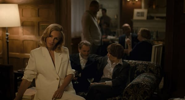 A scene from the film 'After The Hunt' featuring Julia Roberts as Alma in a stylish white suit, seated in a dimly lit gathering with other characters in an elegant setting. The atmosphere is intimate, with guests socializing in the background.