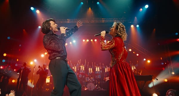A scene from the musical film 'Song Sung Blue' featuring Hugh Jackman as Mike Sardina and Kate Hudson as Claire Stengl singing on stage. They are dressed in glamorous outfits while an enthusiastic audience and band are visible in the background, with vibrant stage lights illuminating the performance.