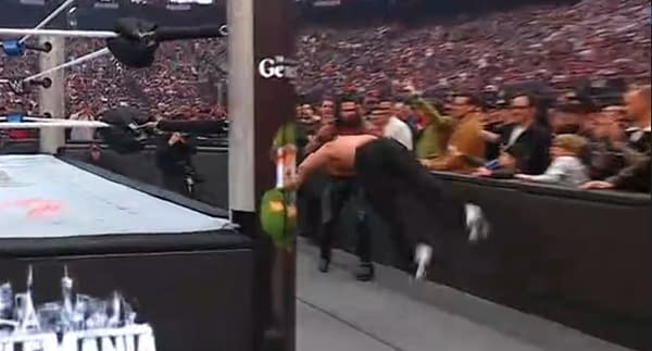 A wrestler executes a spear maneuver, diving toward an opponent at ringside during a wrestling match. The crowd is excited, with fans visible in the background cheering.