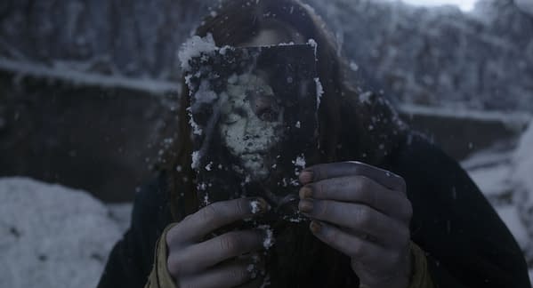 A scene from the movie 'Frankenstein' featuring a figure holding a weathered photograph that shows a distorted face, with snow falling around them. The character, portrayed by Jacob Elordi as The Creature, appears somber amid a bleak, snowy landscape.