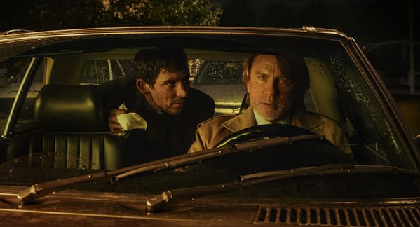 A dramatic scene from the film 'Wake Up Dead Man' featuring two men inside a car. One man, seated in the driver's seat, looks intense and focused, while the other man leans in from the back seat, appearing tense and eager.