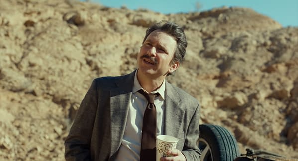 A man with a mustache, dressed in a suit and tie, stands outdoors holding a coffee cup and looking upward, against a backdrop of rocky terrain.