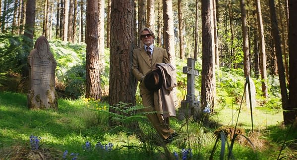 A still from 'Wake Up Dead Man: A Knives Out Mystery,' featuring a man in a tan suit and sunglasses leaning against a tree in a graveyard, surrounded by gravestones and lush greenery.