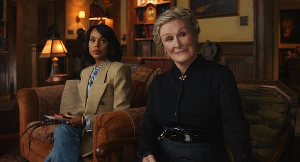 In a scene from 'Wake Up Dead Man: A Knives Out Mystery,' two women sit comfortably on a sofa in a warmly lit room, with a lamp glowing beside them. Kerry Washington, dressed in a light blazer and jeans, holds a notepad, while Glenn Close, in a dark outfit, smiles gently, contributing to the film's intriguing atmosphere.