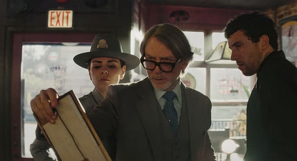 Three characters from 'Wake Up Dead Man: A Knives Out Mystery' are in a bar setting, with a police officer and two men, one examining an object closely. The scene is lit with warm colors, enhancing the tense atmosphere of the moment.