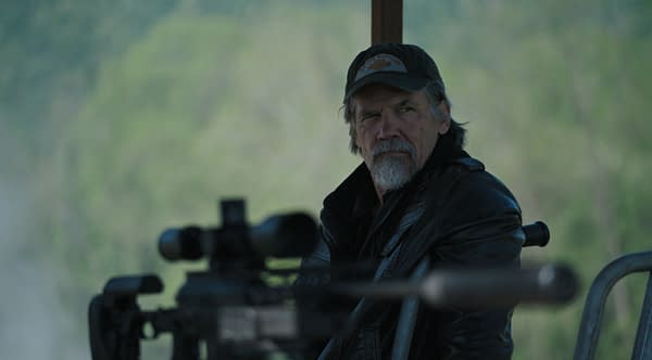 Josh Brolin as Bangley in The Dog Stars, wearing a dark cap and leather jacket while seated beside a rifle in a muted outdoor scene.