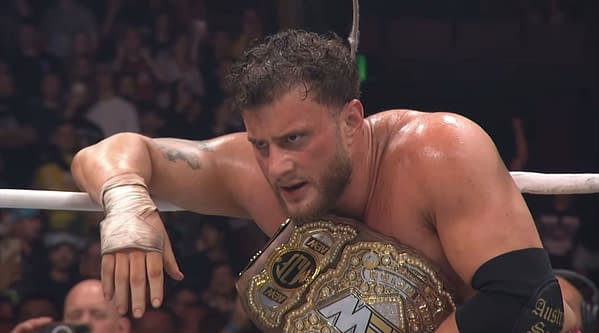 A wrestler, wearing a championship belt and bandaged hand, looks intensely at the audience from the ring at AEW Grand Slam Australia. His sweat glistens as the crowd cheers behind him.