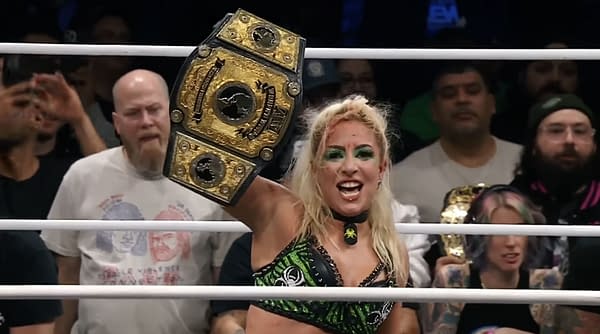 A female wrestler joyfully holds up the AEW Women's World Championship belt, wearing a green and black outfit. The audience is visible in the background, with various expressions of excitement.
