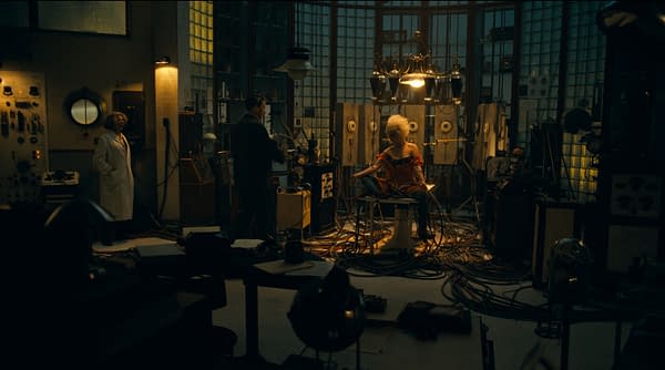 A dramatic scene featuring a woman with curly hair wearing a red dress, sitting in a vintage laboratory surrounded by machinery and tangled wires. A man in a suit stands nearby, with another person in a lab coat observing the situation.