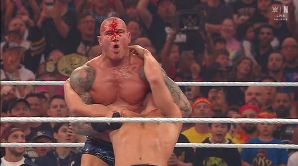 A wrestler with a muscular build and blood on his face is grappling another wrestler in a wrestling ring. The audience in the background shows excitement during the match.