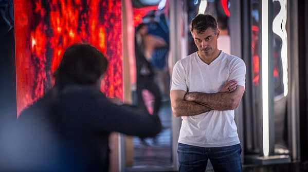 A man in a white t-shirt stands with arms crossed, looking intently at another person off-camera. The background features brightly lit, colorful displays creating a dynamic atmosphere.