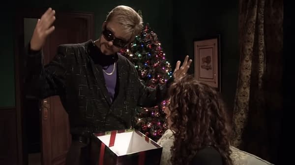 A man in sunglasses and a patterned black jacket gestures dramatically while holding an open gift box, with a Christmas tree lit up in the background. An intrigued woman with curly hair watches.