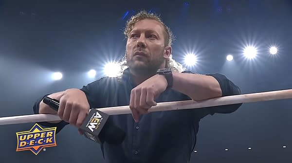 Kenny Omega stands in the AEW ring, leaning on the ropes under bright arena lights and looking serious.