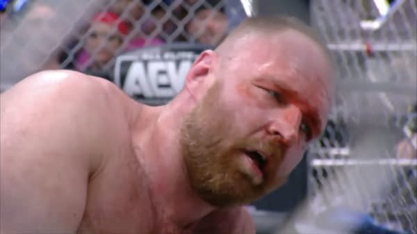 Jon Moxley shows a pained expression after winning a steel cage match against Kenny Omega, with visible wounds on his face and a backdrop of the cage.