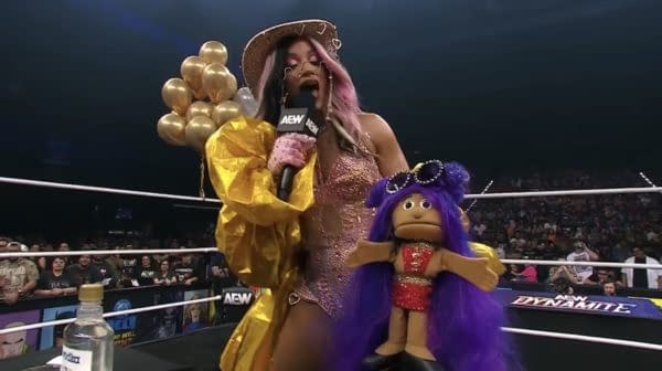 Mercedes Moné stands in the wrestling ring holding a microphone and a colorful puppet, Mini Moné, while wearing a sparkling outfit and surrounded by balloons.