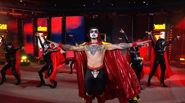 A man in a vibrant red and yellow superhero cape stands center stage with dramatic face paint and tattoos, surrounded by dancers in black costumes with red capes. The backdrop features colorful lights creating an electrifying atmosphere.