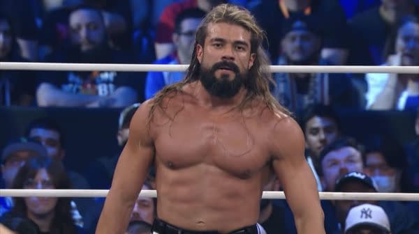 Andrade El Ídolo confidently strides around the wrestling ring during his match, showcasing an intense expression while his muscular physique is visible. Spectators in the background are watching attentively, creating an energetic atmosphere.