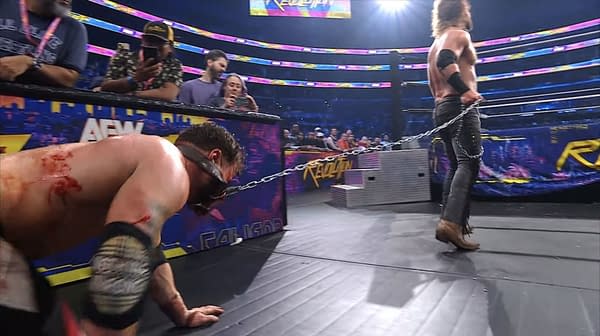 In a wrestling ring, Hangman Adam Page drags AEW World Champion MJF on a chain while MJF crawls on the mat, visibly injured and bloodied. Spectators in the background capture the moment on their phones.