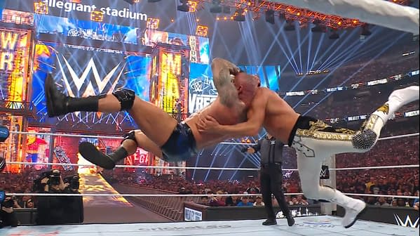 A wrestler executes an RKO move during a match, lifting an opponent into the air with a dramatic flair in a large wrestling arena with bright stage lighting.