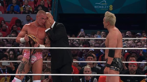 Don Callis leans in to calm Kyle Fletcher as Kazuchika Okada watches during an AEW All In London International Championship triple threat segment at Wembley. The crowd looks on from the arena seats behind the ring.