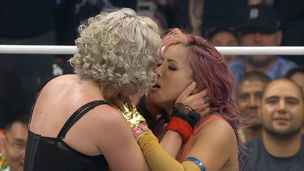 Toni Storm and Mina Shirakawa share a respectful kiss in the wrestling ring, showcasing a moment of sportsmanship after their match. Both wrestlers have distinct hairstyles and colorful wrestling attire.