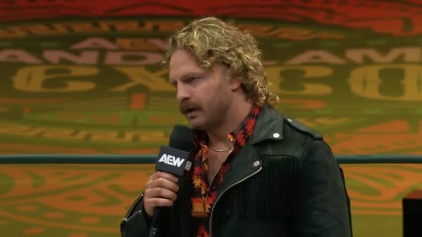 Professional wrestler Hangman Adam Page delivers a passionate promo in Spanish, wearing a colorful shirt and a black jacket with fringes, speaking into a microphone at Arena México for AEW's Grand Slam Mexico event.