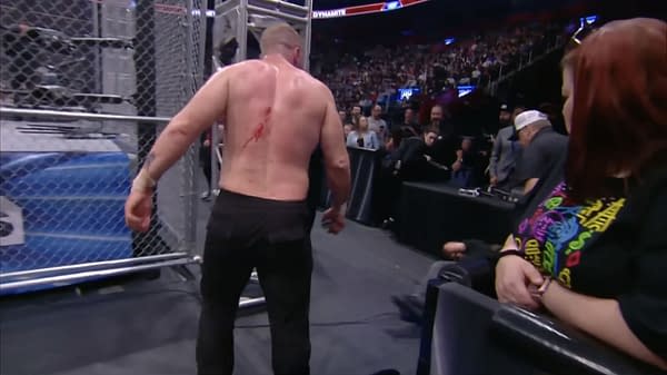 Jon Moxley, with blood on his back, is walking outside the ring during a steel cage wrestling match while a seated crew member is on the ground nearby, amidst a lively crowd during an AEW Dynamite episode.